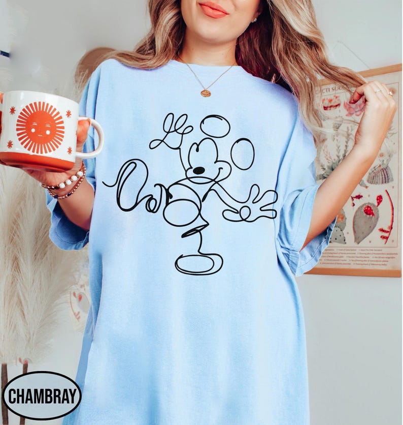 May include: Light blue t-shirt featuring a black line drawing of Mickey Mouse with the word "Cool" written in cursive. A white and orange mug with a sun design is held in the left hand. The word "Chambray" is printed on the bottom left.