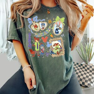 May include: Dark green t-shirt featuring characters from the movie Zootopia. The shirt has a colorful graphic design with framed images of the characters, the word "Zootopia", and Mickey Mouse balloons.