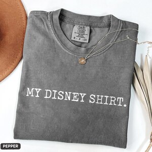 May include: A gray t-shirt with the text "MY DISNEY SHIRT." in white. The shirt has a crew neck and short sleeves. A silver necklace with a pendant is draped across the shirt. The shirt is folded and has a tag that says "COMFORT COLORS."