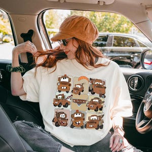 Może przedstawiać: Kremowa koszulka z nadrukiem w kratkę z postaciami z kresk&oacute;wek samochod&oacute;w w odcieniach brązu i beżu. Na koszulce nadrukowano napis "Cars". Osoba na zdjęciu ma na sobie brązowy sztruksowy kapelusz i okulary przeciwsłoneczne.