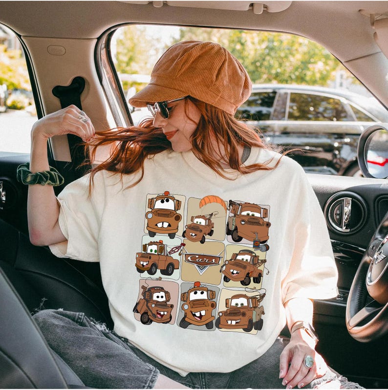 Może przedstawiać: Kremowa koszulka z nadrukiem w kratkę z postaciami z kresk&oacute;wek samochod&oacute;w w odcieniach brązu i beżu. Na koszulce nadrukowano napis "Cars". Osoba na zdjęciu ma na sobie brązowy sztruksowy kapelusz i okulary przeciwsłoneczne.