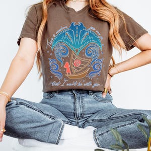 May include: Brown t-shirt featuring a graphic design with a boat, a figure, and swirling blue and teal elements. The text on the shirt reads "There is nowhere you are that I won't be with you..."