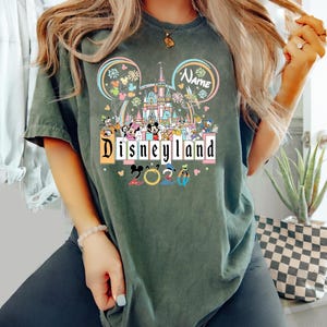 Op de afbeelding: Donkergroen t-shirt met een Disneyland-afbeelding. Het ontwerp toont het Disneyland-logo, een kasteel, Mickey Mouse-oren en verschillende Disney-personages. Het jaar "2023" is ook in het ontwerp opgenomen.