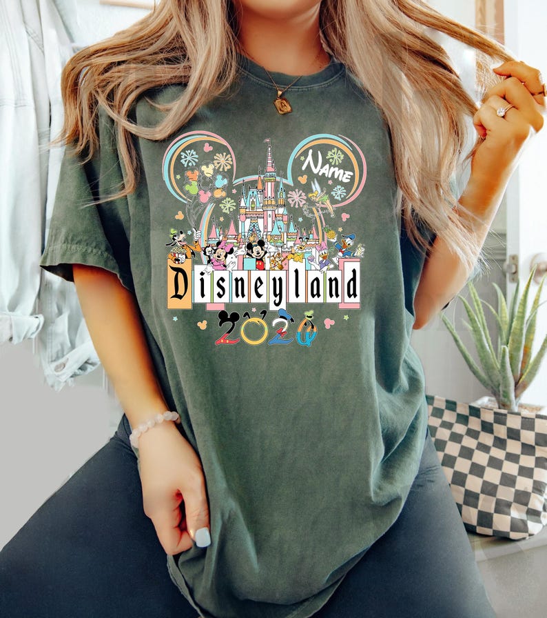 Op de afbeelding: Donkergroen t-shirt met een Disneyland-afbeelding. Het ontwerp toont het Disneyland-logo, een kasteel, Mickey Mouse-oren en verschillende Disney-personages. Het jaar "2023" is ook in het ontwerp opgenomen.