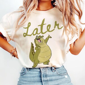 May include: Cream-colored t-shirt featuring a cartoon alligator with its arms raised. The word "Later" is written above the alligator in a green, cursive font. The alligator is green with a distressed design.