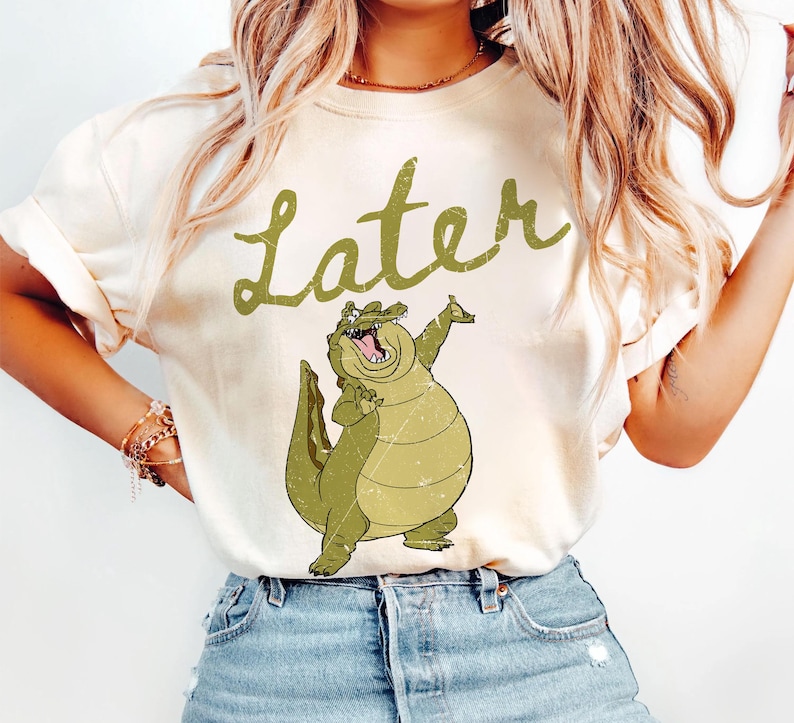 May include: Cream-colored t-shirt featuring a cartoon alligator with its arms raised. The word "Later" is written above the alligator in a green, cursive font. The alligator is green with a distressed design.