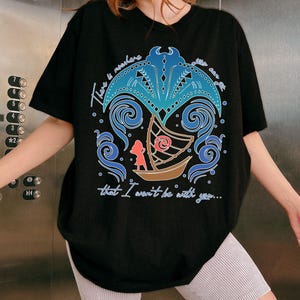 May include: Black t-shirt featuring a colorful graphic design with the text "There is nowhere that I won't be with you...". The design includes a boat, a figure, and stylized waves and palm tree elements in blue and brown.