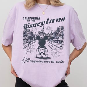 May include: A light purple t-shirt with a graphic of Mickey Mouse in front of Disneyland. The text on the shirt reads "CALIFORNIA Est. 1955 Disneyland" and "The happiest place on earth."