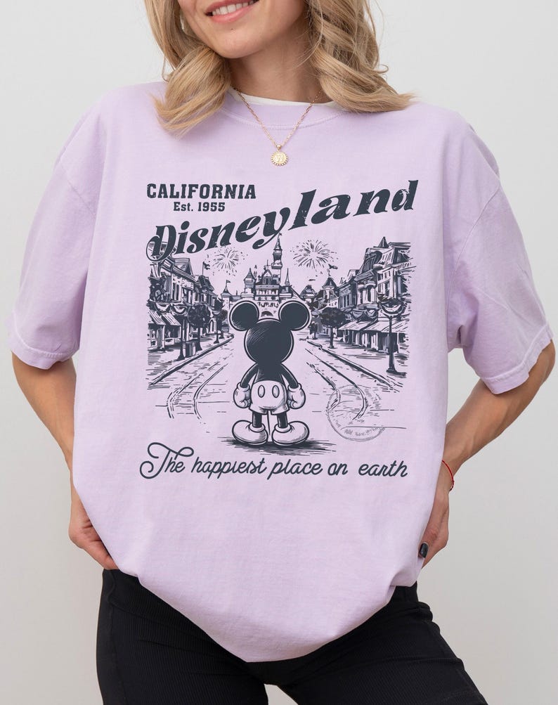 May include: A light purple t-shirt with a graphic of Mickey Mouse in front of Disneyland. The text on the shirt reads "CALIFORNIA Est. 1955 Disneyland" and "The happiest place on earth."