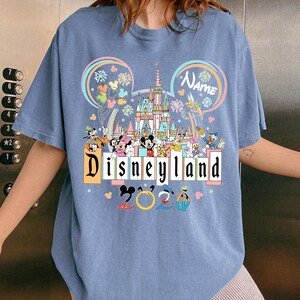 Op de afbeelding: Lichtblauw T-shirt met een kleurrijke Disneyland-afbeelding. Het ontwerp bevat het Disneyland-logo, een kasteel, Mickey Mouse-oren en verschillende Disney-personages. De tekst "2023" is ook aanwezig.