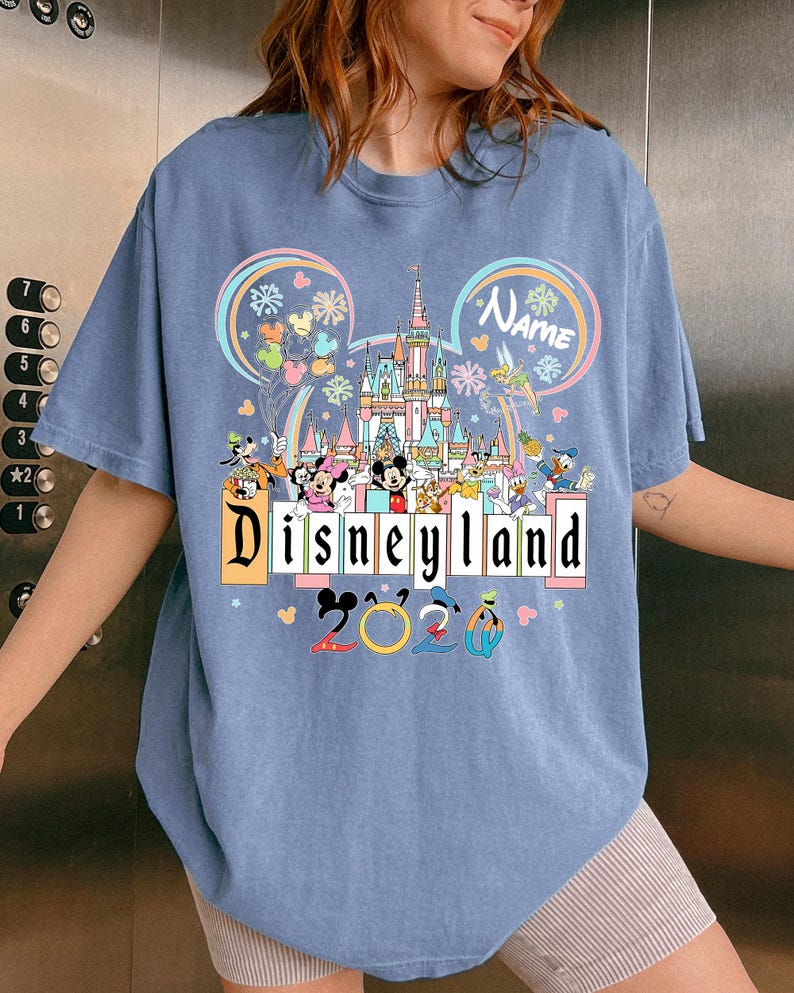 Op de afbeelding: Lichtblauw T-shirt met een kleurrijke Disneyland-afbeelding. Het ontwerp bevat het Disneyland-logo, een kasteel, Mickey Mouse-oren en verschillende Disney-personages. De tekst "2023" is ook aanwezig.