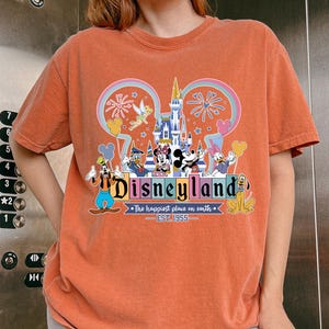 Może przedstawiać: Pomarańczowa koszulka z kolorową grafiką Disneyland. Projekt zawiera logo Disneyland, Myszka Miki i napis "The happiest place on earth. EST. 1955."