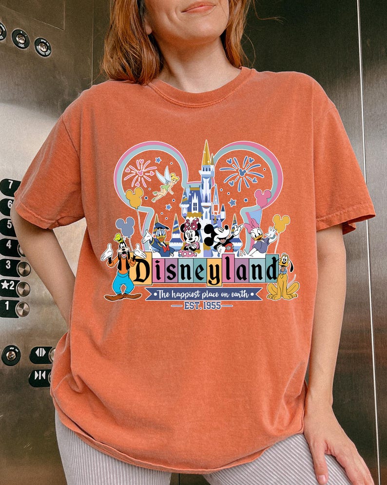 Może przedstawiać: Pomarańczowa koszulka z kolorową grafiką Disneyland. Projekt zawiera logo Disneyland, Myszka Miki i napis "The happiest place on earth. EST. 1955."