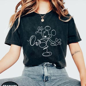 May include: Graphite-colored t-shirt featuring a white line drawing of Mickey Mouse. The design includes the character's outline and the word "Oh" in a cursive font. The shirt is paired with blue denim jeans.