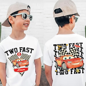 May include: White t-shirt featuring a cartoon race car design. The front of the shirt says "TWO FAST" with a number 2 and a name. The back of the shirt has the same design. The shirt is worn by a child.
