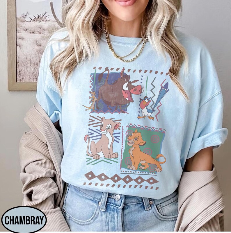 May include: Light blue t-shirt featuring a colorful graphic of characters from The Lion King. The design includes images of Timon, Pumbaa, Simba, and Nala, set against a geometric background. The shirt has a relaxed fit and short sleeves.