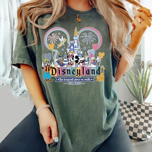 Może przedstawiać: Ciemnozielona koszulka z kolorową grafiką Disneylandu. Projekt zawiera zamek, fajerwerki i r&oacute;żne postacie Disneya. Tekst "Disneyland" jest wyraźnie wyświetlany, wraz z "The happiest place on earth - EST. 1955".