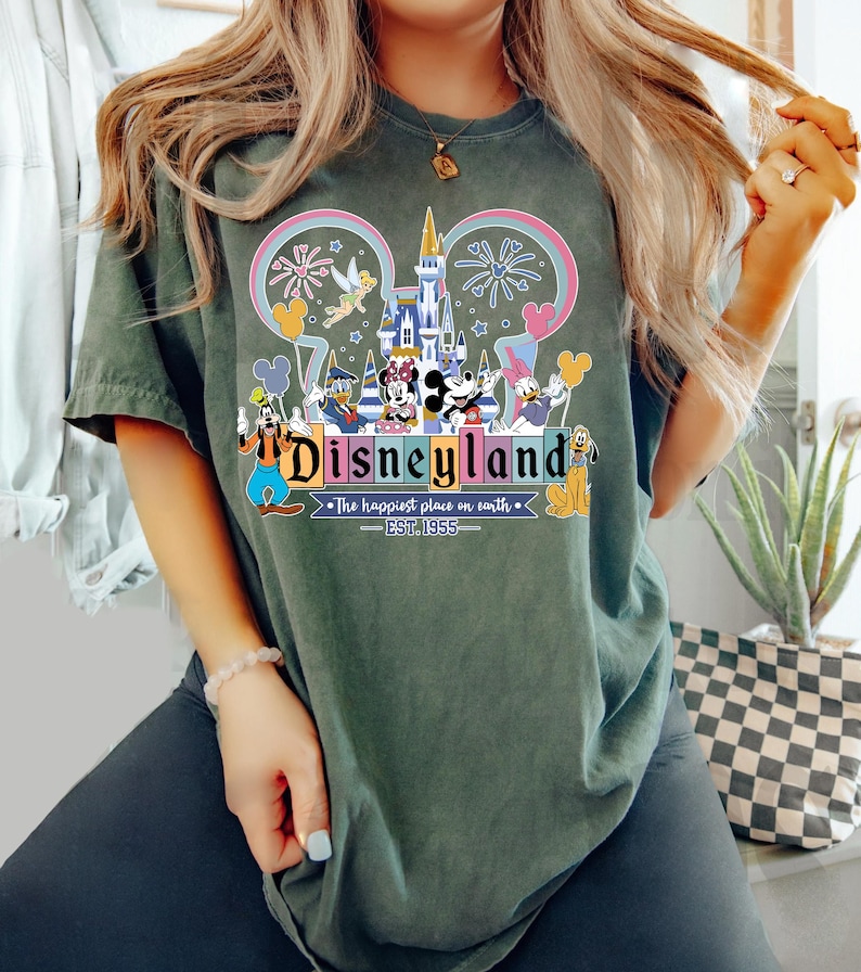 Może przedstawiać: Ciemnozielona koszulka z kolorową grafiką Disneylandu. Projekt zawiera zamek, fajerwerki i r&oacute;żne postacie Disneya. Tekst "Disneyland" jest wyraźnie wyświetlany, wraz z "The happiest place on earth - EST. 1955".