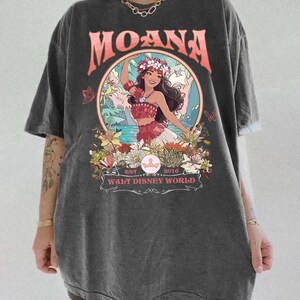 May include: Dark gray t-shirt featuring a colorful graphic of Moana from Walt Disney World. The design includes the word "MOANA" in coral letters above a circular image of Moana, surrounded by flowers and butterflies. The text "EST 2016" and "WALT DISNEY WORLD" are also included.