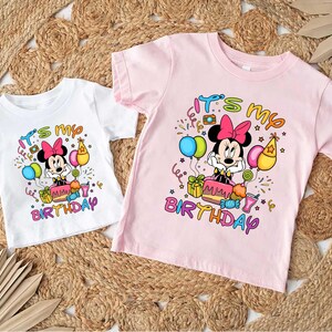 May include: Two short-sleeved t-shirts, one white and one pink, featuring a Minnie Mouse graphic. The graphic includes the text "It's my Birthday" with colorful balloons, confetti, and a birthday cake. The shirts are likely for children.