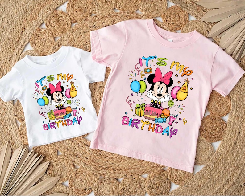 May include: Two short-sleeved t-shirts, one white and one pink, featuring a Minnie Mouse graphic. The graphic includes the text "It's my Birthday" with colorful balloons, confetti, and a birthday cake. The shirts are likely for children.