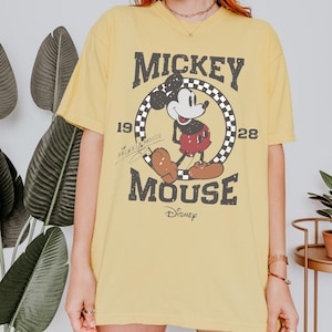 May include: Yellow t-shirt featuring a distressed graphic of Mickey Mouse. The design includes the text "Mickey Mouse" and the years "1928" and "19". The Disney logo is at the bottom. The shirt is a casual, oversized fit.