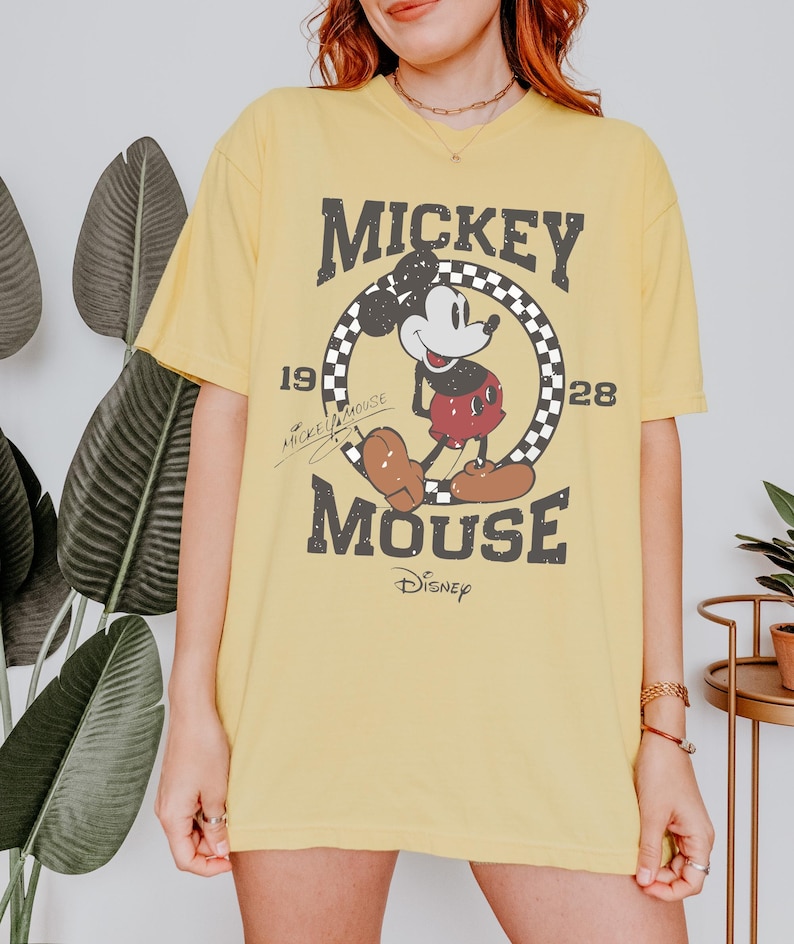 May include: Yellow t-shirt featuring a distressed graphic of Mickey Mouse. The design includes the text "Mickey Mouse" and the years "1928" and "19". The Disney logo is at the bottom. The shirt is a casual, oversized fit.