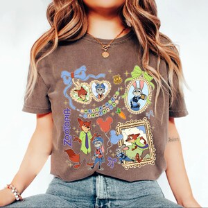 May include: Brown t-shirt featuring colorful illustrations of characters from the animated film "Zootopia." The design includes framed portraits, character figures, and the film's title. The shirt has a crew neck and short sleeves.