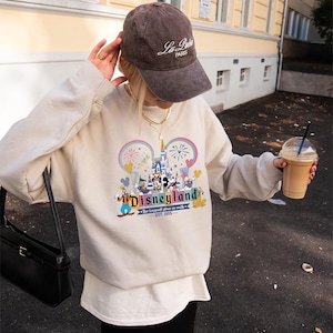 Może przedstawiać: Beżowa bluza z nadrukiem Disneyland z Myszką Miki i przyjaci&oacute;łmi. Tekst "Disneyland" jest wyświetlany w fantazyjnej czcionce, z frazą "The happiest place on earth" poniżej. Widoczna jest r&oacute;wnież brązowa czapka z daszkiem z napisem "La Paris".