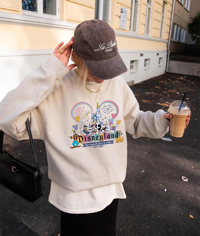 Może przedstawiać: Beżowa bluza z nadrukiem Disneyland z Myszką Miki i przyjaci&oacute;łmi. Tekst "Disneyland" jest wyświetlany w fantazyjnej czcionce, z frazą "The happiest place on earth" poniżej. Widoczna jest r&oacute;wnież brązowa czapka z daszkiem z napisem "La Paris".