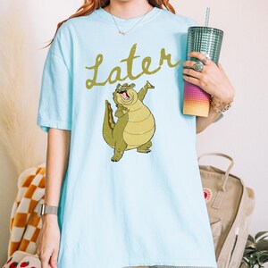 May include: Light blue t-shirt featuring a cartoon alligator with the word "Later" in gold script. The alligator is green and yellow. The shirt is worn with gray shorts and a watch.