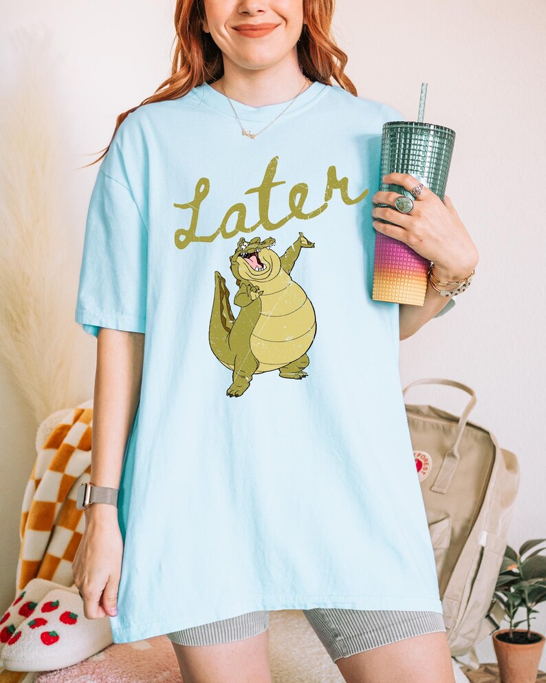 May include: Light blue t-shirt featuring a cartoon alligator with the word "Later" in gold script. The alligator is green and yellow. The shirt is worn with gray shorts and a watch.