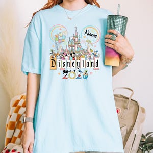 Op de afbeelding: Lichtblauw T-shirt met een Disneyland-ontwerp met de tekst "Disneyland 2020". Het ontwerp bevat een kasteel, stripfiguren en de naam "Name" in een cirkel. Het shirt heeft korte mouwen.