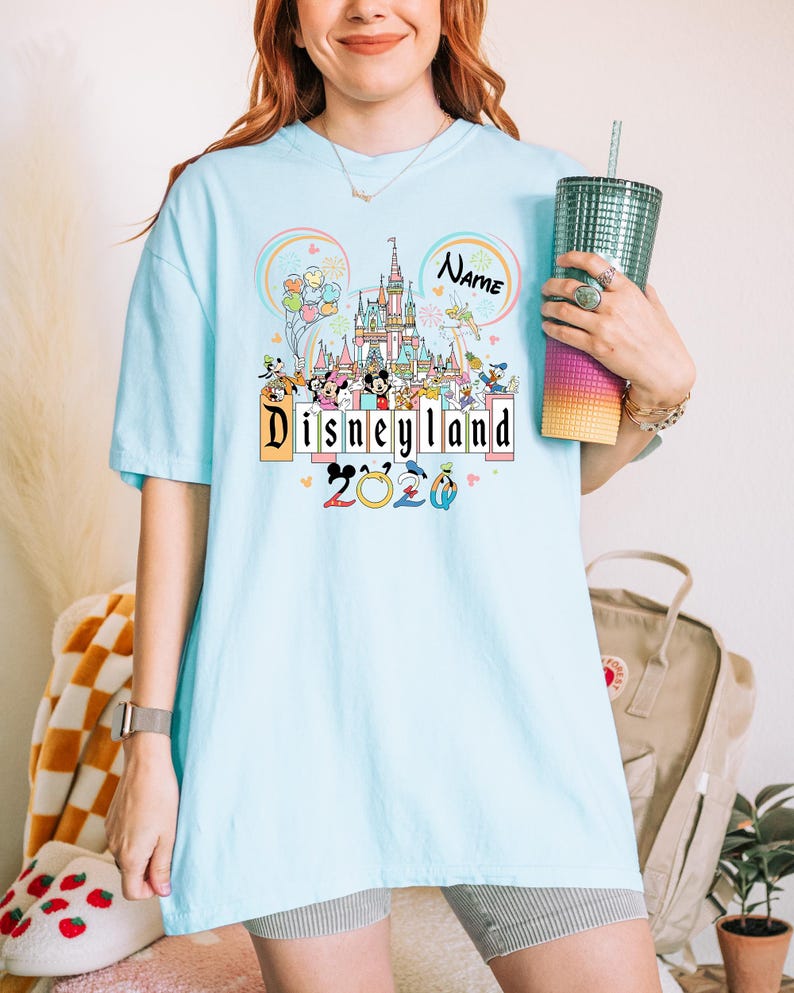 Op de afbeelding: Lichtblauw T-shirt met een Disneyland-ontwerp met de tekst "Disneyland 2020". Het ontwerp bevat een kasteel, stripfiguren en de naam "Name" in een cirkel. Het shirt heeft korte mouwen.