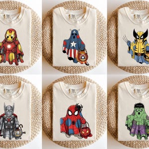 May include: Six folded cream-colored t-shirts with superhero ghost designs. Each shirt features a different character: Iron Man, Captain America, Wolverine, Thor, Spider-Man, and the Hulk, each holding a Halloween pumpkin bucket.