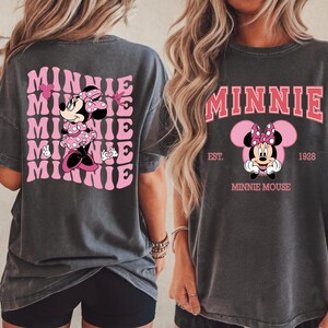 May include: Dark gray t-shirts featuring Minnie Mouse. One shirt has a pink and white design with the word "Minnie" repeated. The other shirt has a Minnie Mouse graphic with the word "Minnie" above and "Minnie Mouse" below.