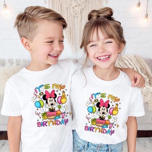 May include: Two children wearing white t-shirts with a colorful Minnie Mouse birthday design. The design includes Minnie Mouse, balloons, a cake, and the text "It's my birthday".