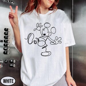 May include: White t-shirt featuring a black line drawing of Mickey Mouse. The design shows Mickey with his arms outstretched and the word "Oh" in cursive. The shirt is a casual style, with short sleeves.