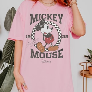 May include: Pink t-shirt featuring a distressed graphic of Mickey Mouse. The design includes Mickey in a circle with a checkered border, the text "MICKEY MOUSE", and the year "1928". The Disney logo is at the bottom.