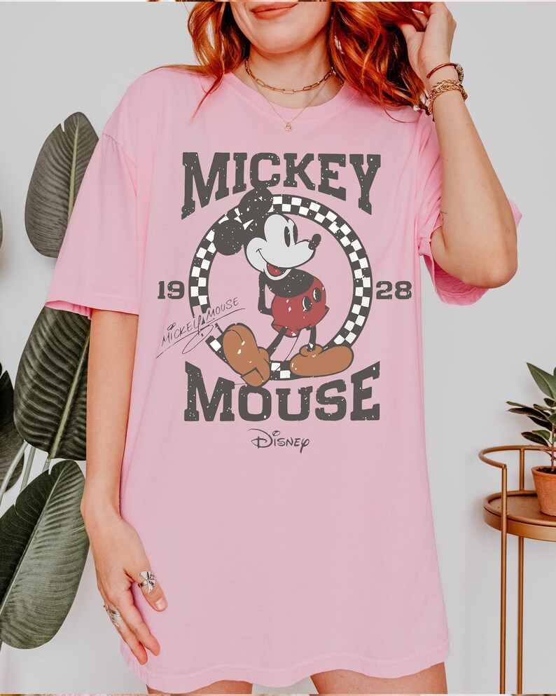 May include: Pink t-shirt featuring a distressed graphic of Mickey Mouse. The design includes Mickey in a circle with a checkered border, the text "MICKEY MOUSE", and the year "1928". The Disney logo is at the bottom.