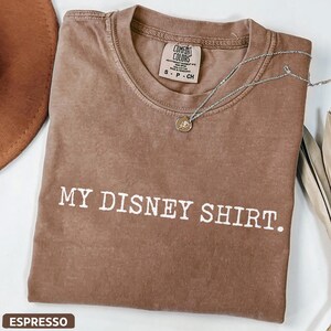 May include: Espresso-colored t-shirt with the text "MY DISNEY SHIRT." A silver necklace with a pendant rests on the shirt. The shirt has a crew neck and a tag that reads "COMFORT COLORS."