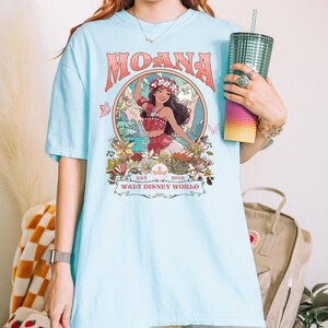May include: Light blue t-shirt featuring the word "MOANA" in a retro font above a graphic of a woman in a floral crown. The graphic is surrounded by flowers and butterflies, with the text "WALT DISNEY WORLD" below.