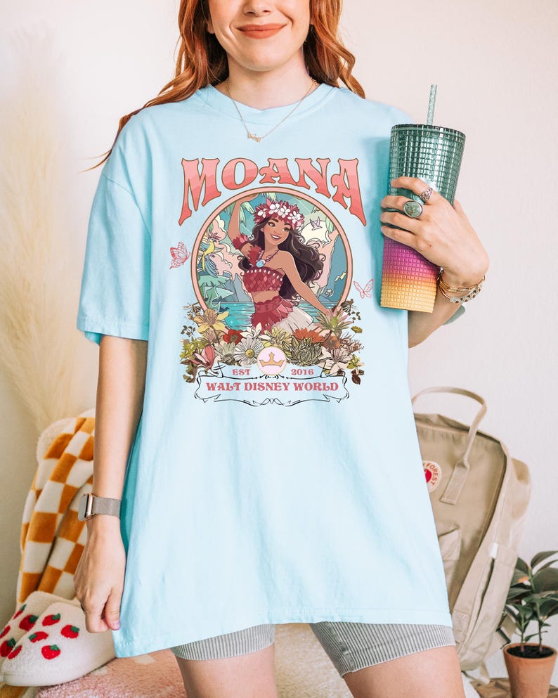 May include: Light blue t-shirt featuring the word "MOANA" in a retro font above a graphic of a woman in a floral crown. The graphic is surrounded by flowers and butterflies, with the text "WALT DISNEY WORLD" below.