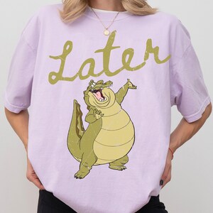 May include: A light purple t-shirt featuring a cartoon alligator with the word "Later" in a script font above it. The alligator is green and yellow, with its mouth open and arms raised. The shirt is a casual style.