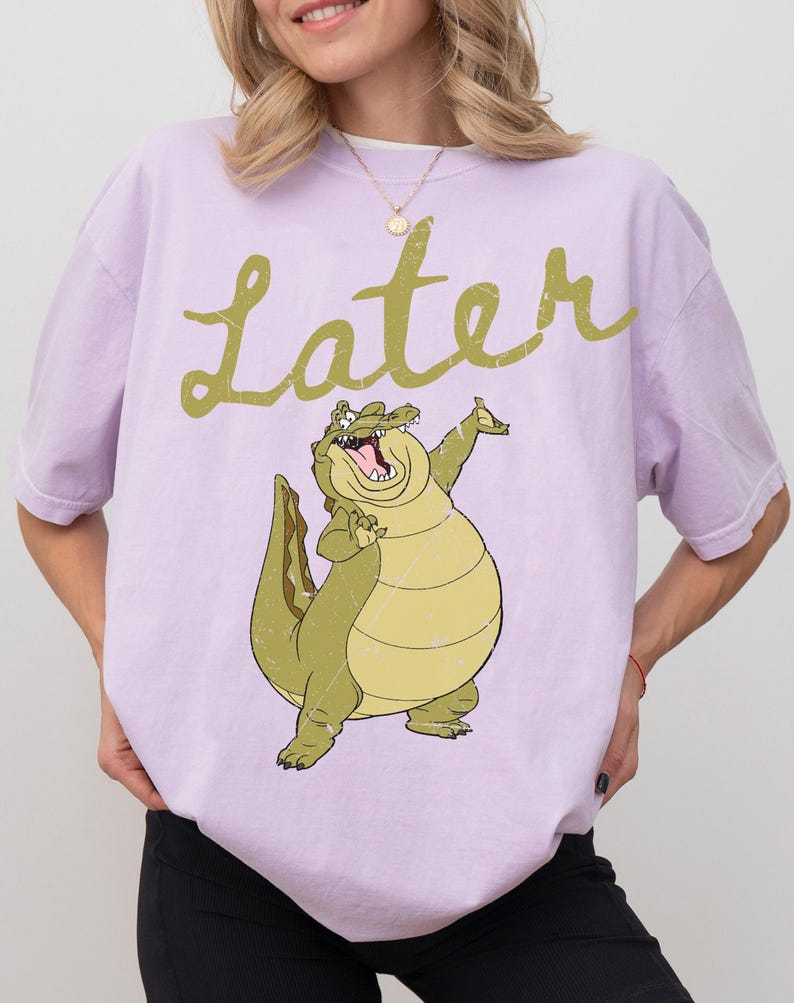 May include: A light purple t-shirt featuring a cartoon alligator with the word "Later" in a script font above it. The alligator is green and yellow, with its mouth open and arms raised. The shirt is a casual style.