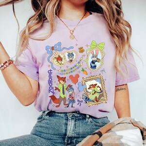 May include: A light purple t-shirt featuring characters from the animated movie Zootopia. The design includes framed images of the characters, along with the word "Zootopia" in a vertical font. The shirt is made of a soft material.