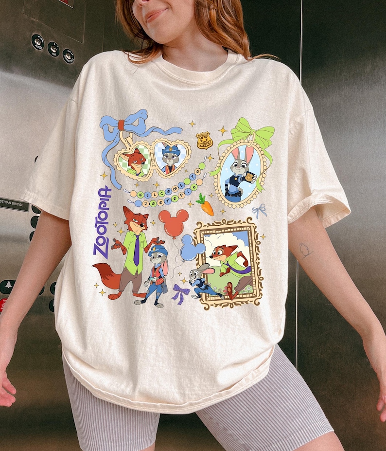 May include: Cream-colored t-shirt featuring a colorful Zootopia graphic. The design includes cartoon characters, frames, and the word "Zootopia" in blue. The shirt has short sleeves and a relaxed fit.