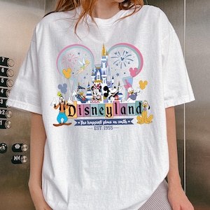 Może przedstawiać: Biała koszulka z kolorową grafiką Disneylandu. Projekt zawiera zamek, Myszka Miki, Myszka Minnie, Kaczor Donald, Goofy i Pluto. Wyświetlany jest r&oacute;wnież tekst "Disneyland" i "The happiest place on earth - EST. 1955".