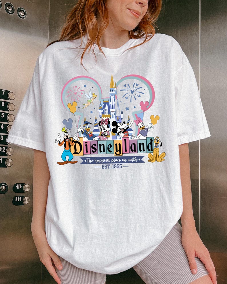 Może przedstawiać: Biała koszulka z kolorową grafiką Disneylandu. Projekt zawiera zamek, Myszka Miki, Myszka Minnie, Kaczor Donald, Goofy i Pluto. Wyświetlany jest r&oacute;wnież tekst "Disneyland" i "The happiest place on earth - EST. 1955".