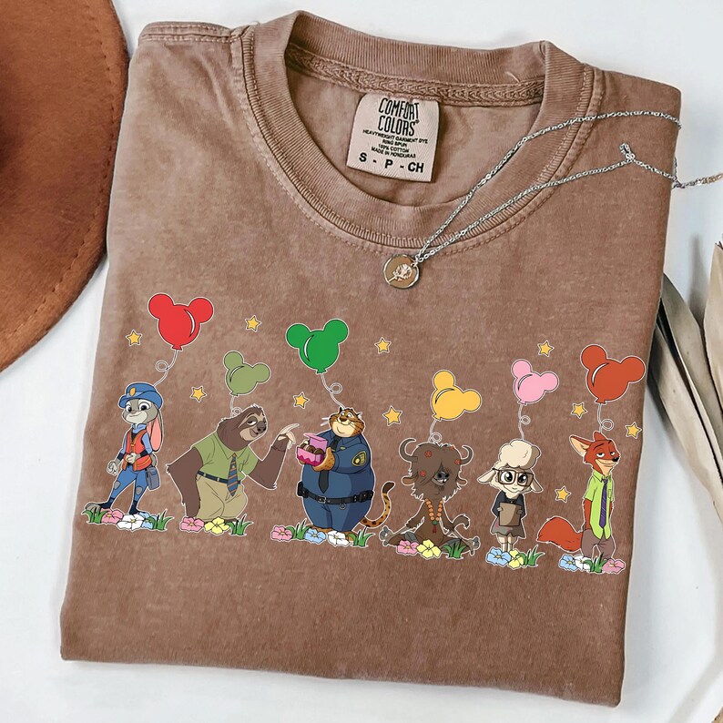 以下が含まれることがあります： 風船を持った漫画のキャラクターが描かれた茶色のコンフォートカラーズTシャツ。キャラクターは花の列に立っています。シルバーのネックレスがシャツにかけられています。シャツには「Comfort Colors」と書かれた小さなタグが付いています。